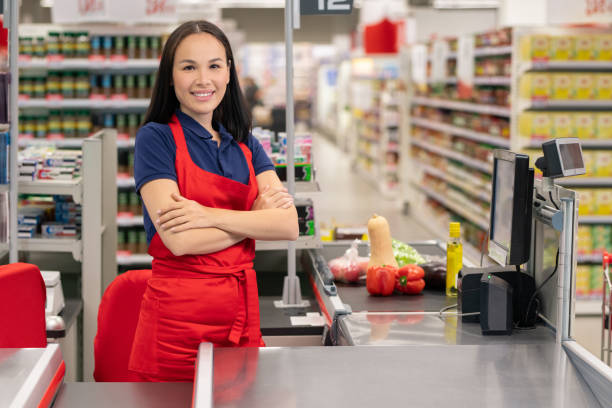 Attractive Asian woman working in supermarket standing at cash desk with arms crossed looking at camera smiling